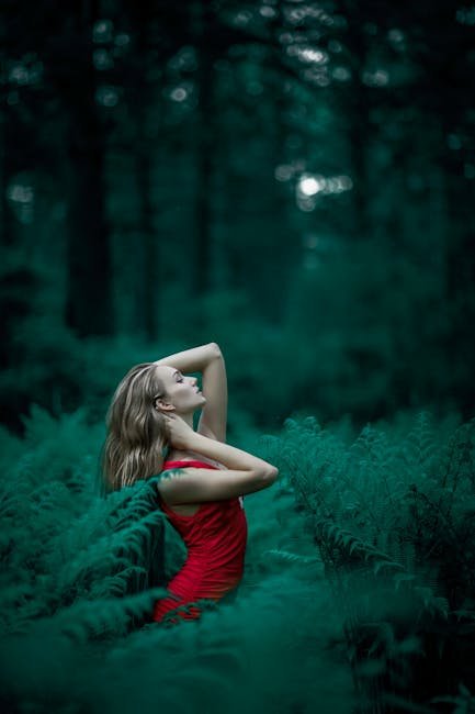 A woman poses in a vibrant red dress amidst lush green ferns in an enchanting forest setting.